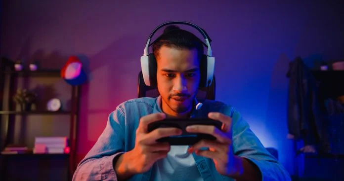 Un joueur asiatique heureux porte un casque de compétition pour jouer à un jeu vidéo en ligne avec un smartphone coloré au néon dans le salon la nuit d'une maison moderne. Comment promouvoir un jeu mobile